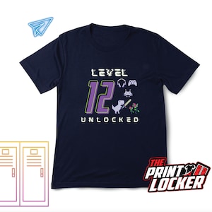 May include: Navy blue t-shirt with a purple graphic that says "Level 12 Unlocked". The graphic also includes a pixelated dinosaur, a video game controller, and a flower.