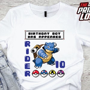 Puede incluir: Camiseta blanca con un personaje pixelado azul y verde, el texto "Birthday Boy Has Appeared" y el texto "Rider 10".
