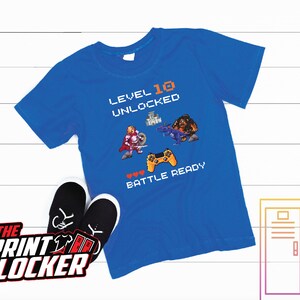 May include: A royal blue t-shirt with a pixelated graphic of a video game character, a castle, and a dragon. The text on the shirt reads "Level 10 Unlocked" and "Battle Ready".