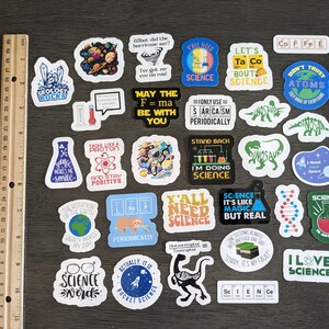Puede incluir: Una colecci&oacute;n de 25 pegatinas con tem&aacute;tica cient&iacute;fica que presentan varios dise&ntilde;os, incluidos dinosaurios, planetas, vasos de precipitados y texto. Las pegatinas est&aacute;n sobre una superficie de madera marr&oacute;n con una regla.