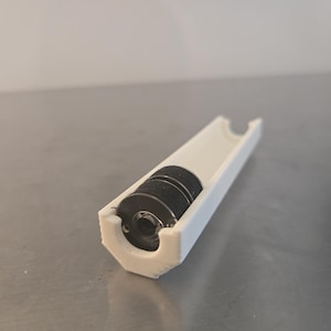 May include: A white, 3D-printed object with a cylindrical cavity containing several black, circular components. The object has a hexagonal end and a channel running along its length. The design suggests a mechanical or technical product.