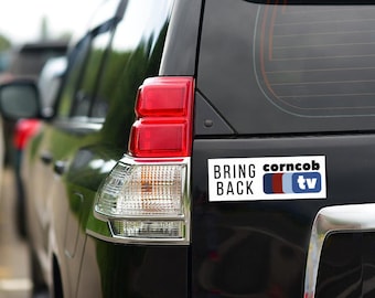 以下が含まれることがあります： 「Bring Back corncob tv」と書かれた白いバンパーステッカーが貼られた黒いSUV。ステッカーには赤、白、青のロゴがあります。