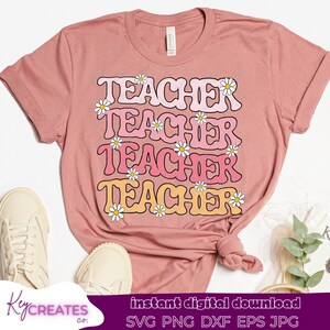 Puede incluir: Una camiseta rosa con la palabra "TEACHER" repetida cuatro veces en una fuente retro con flores de margarita alrededor de cada palabra. La camiseta está doblada y se muestra sobre una superficie blanca con zapatos blancos.