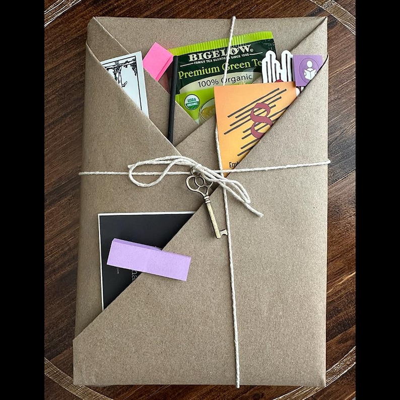 May include: A brown paper-wrapped parcel tied with string, adorned with a small gold key charm. The package includes a green tea bag, a pink sticky note, and a small orange card. The overall aesthetic is rustic and gift-like.