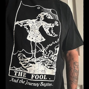 May include: Black t-shirt with a white graphic of a tarot card, The Fool. The card depicts a figure walking towards a cliff with a staff and a backpack. The text "The Fool. And the Journey Begins..." is printed below the card.