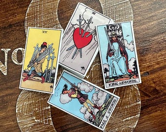 May include: A collection of tarot cards featuring colorful illustrations. The cards depict various scenes, including a heart pierced by swords, a figure holding swords, and a king with a sword. The cards are arranged on a wooden surface.