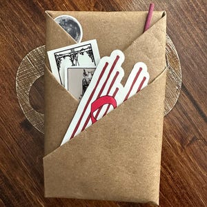 May include: A brown paper package with a red and white striped sticker, a tarot card, and a small moon sticker. The package is folded into a triangle shape.