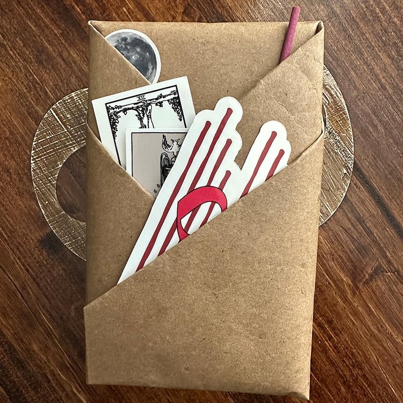 May include: A brown paper package with a red and white striped sticker, a tarot card, and a small moon sticker. The package is folded into a triangle shape.