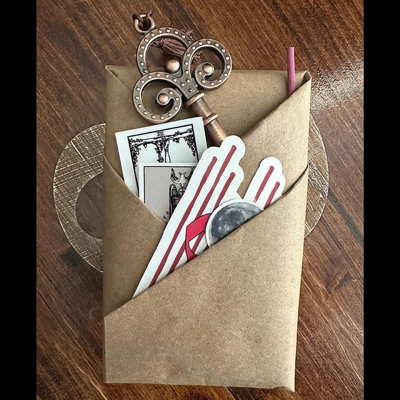 May include: A brown paper envelope with a decorative key, a tarot card, and a sticker with a red and white striped design and a moon.