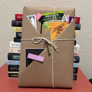May include: A brown paper-wrapped gift tied with string, with a small key charm. The package contains a green tea packet, cards, and a stack of books in the background. A pink sticky note is attached to the front.