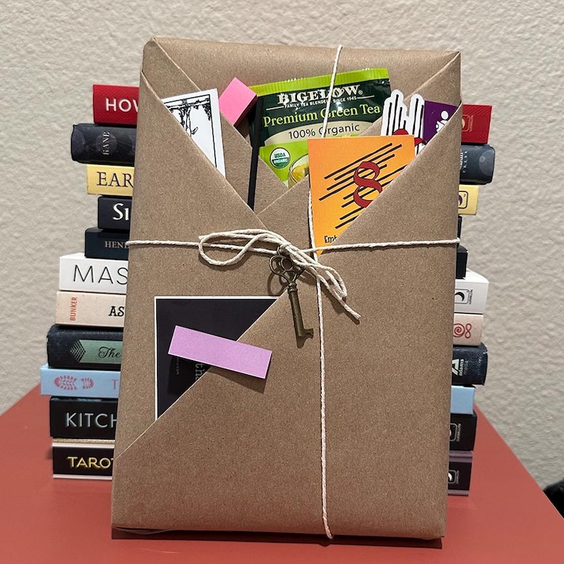 May include: A brown paper-wrapped gift tied with string, with a small key charm. The package contains a green tea packet, cards, and a stack of books in the background. A pink sticky note is attached to the front.