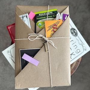 May include: A parcel wrapped in brown paper, secured with twine and a decorative key. It contains tea, cards, and a pink sticky note. The package rests on a neutral surface.