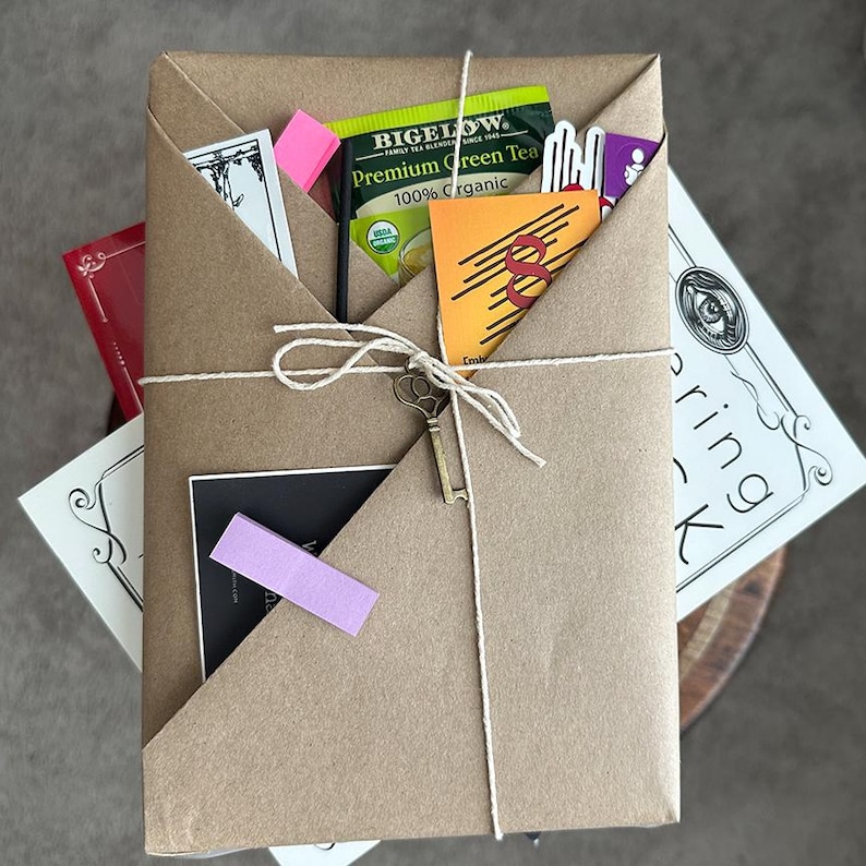 May include: A parcel wrapped in brown paper, secured with twine and a decorative key. It contains tea, cards, and a pink sticky note. The package rests on a neutral surface.