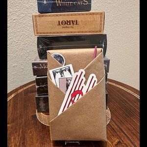 May include: A brown paper envelope with a red and white striped sticker, a black and white sticker of a tarot card, and a small white sticker with a moon design. The envelope is sitting on top of a stack of tarot card boxes.