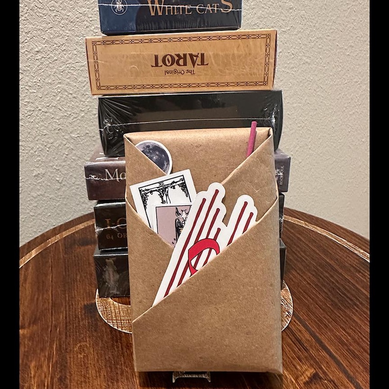 May include: A brown paper envelope with a red and white striped sticker, a black and white sticker of a tarot card, and a small white sticker with a moon design. The envelope is sitting on top of a stack of tarot card boxes.