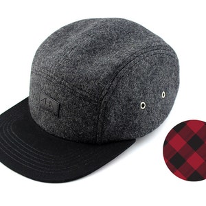 May include: A gray wool 5-panel cap with a black brim and a black leather patch with the letters "AA" on the front. The cap has a red and black plaid pattern on the back.