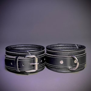 Op de afbeelding: Een paar zwarte leren manchetten met zilveren hardware. Een manchet heeft een gespsluiting en de andere een ring.