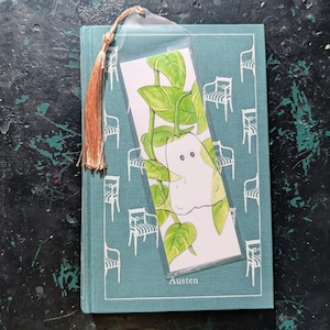 May include: A clear plastic bookmark with a watercolor illustration of a white ghost-like figure with green leaves. The bookmark has a brown tassel and is placed on a teal book with the name "Austen" on the cover.