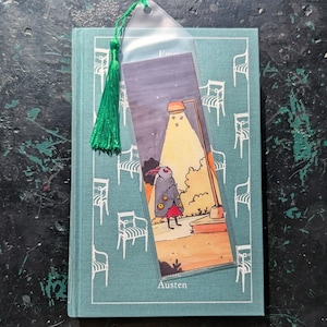 May include: A teal book with a white chair pattern and the name "Austen" printed on the bottom. A bookmark with a cartoon moth under a street light is inserted. A green tassel hangs from the bookmark.