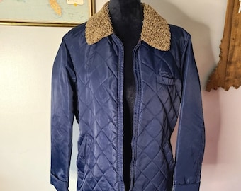 May include: A navy blue quilted jacket with a tan faux fur collar. The jacket has a diamond pattern, a front zipper, and two front pockets. The jacket is displayed on a black mannequin.