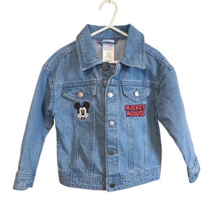Op de afbeelding: Lichtblauwe spijkerjas met een klassieke kraag en knoopsluiting. Voorzien van een Mickey Mouse-patch op de borst en een "Mickey Mouse"-patch aan de rechterkant. De jas hangt aan een witte hanger.