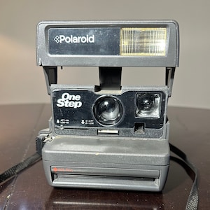 Peut inclure: Un appareil photo instantané Polaroid One Step noir avec un flash intégré. L'appareil photo est doté d'une sangle noire.