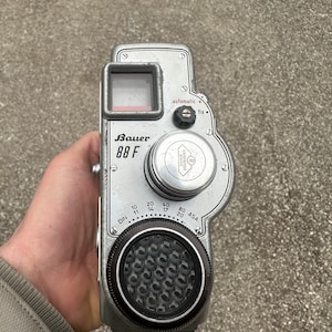 May include: A silver Bauer 88 F film camera with a black lens and a silver dial with markings for DIN and ASA. The camera has a built-in light meter and a viewfinder.
