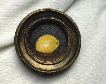 May include: A miniature framed painting of a lemon. The artwork features a yellow lemon against a dark brown backdrop, enclosed within a circular, gold-toned frame. The painting is a small still life, capturing the fruit in a simple, elegant style.