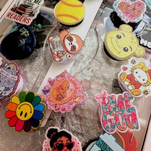 May include: A collection of various circular phone grips featuring diverse designs including a yellow softball, a sloth holding a coffee mug, a rainbow flower, a Winnie the Pooh illustration, a floral heart, and text-based graphics like Take Me To Your Readers.