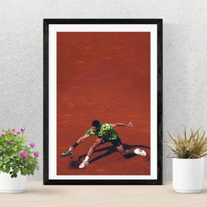 May include: A framed poster of a tennis player in a green and black shirt, diving to hit a tennis ball on a red clay court.