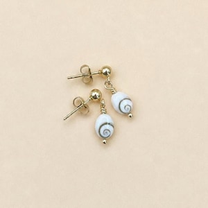 May include: A pair of gold stud earrings featuring dangling white shiva shell beads. Each bead is oval-shaped with a natural grey spiral pattern on the front. The earrings are constructed with gold posts and small gold wire loops connecting the beads.