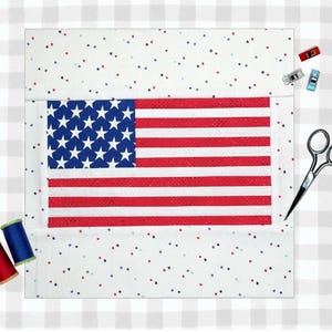Pode incluir: Um quadrado de tecido com o desenho da bandeira americana, com listras vermelhas e brancas e estrelas azuis. O tecido é cercado por uma borda branca com manchas vermelhas, azuis e pretas. Materiais de costura, incluindo tesouras, carretéis de linha e clipes, são dispostos ao redor do tecido.