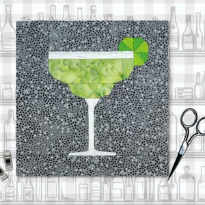 May include: A fabric art piece featuring a margarita cocktail. The drink is green with a white rim, and a lime wedge. The background is gray with black dots. A pair of scissors and clips are in the image.
