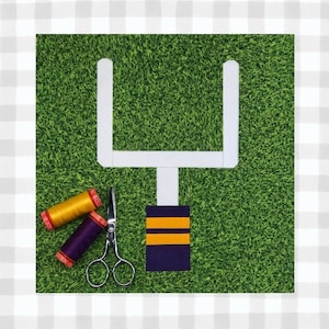 May include: A white felt football goalpost with a purple and yellow felt stripe on the base. There are two spools of thread, one yellow and one purple, and a pair of silver scissors on a green felt background.