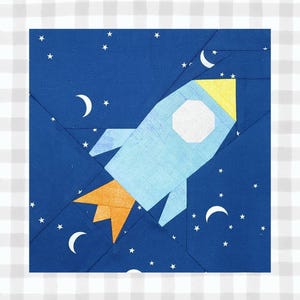 May include: A square fabric block featuring a light blue rocket ship with yellow and orange accents, set against a dark blue background with white stars and crescent moons. The rocket has a white octagon window.