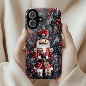 Może przedstawiać: Etui na telefon z świątecznym motywem Dziadka do Orzechów. Etui ma ciemne tło z czerwonymi jagodami i białymi igłami sosnowymi. Dziadek do Orzechów ubrany jest w czerwono-czarny mundur i trzyma złoty kij.