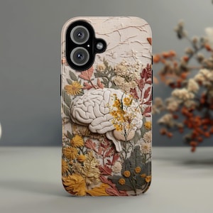 May include: A phone case featuring a 3D design of a brain with floral accents in yellow, orange, and cream colors. The case has a textured, off-white background and a dual-lens camera cutout.