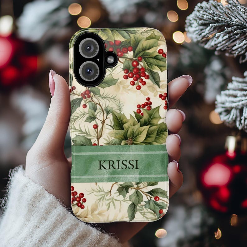 May include: A phone case featuring a festive design with green leaves, red berries, and a cream-colored background. A green banner at the bottom displays the name "KRISSI". The case has a camera cutout and is held in a hand.