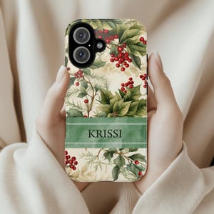 May include: A phone case featuring a botanical design with green leaves, red berries, and the name "KRISSI" on a green banner. The case has a cream-colored background and a camera cutout.
