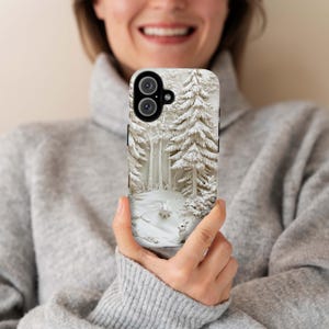 May include: A white phone case featuring a winter scene with a snowy forest and two white rabbits. The case has a black camera lens cutout.