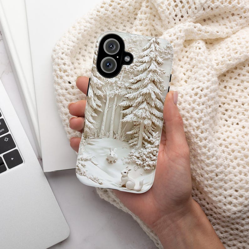 May include: White phone case featuring a 3D design of a snowy woodland scene with two white rabbits. The case has a black camera lens cutout.