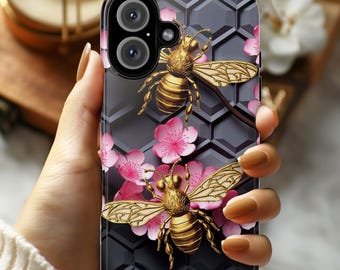May include: A black phone case with a honeycomb pattern and two bees with pink flowers. The bees are gold and have black eyes. The flowers are pink and white.