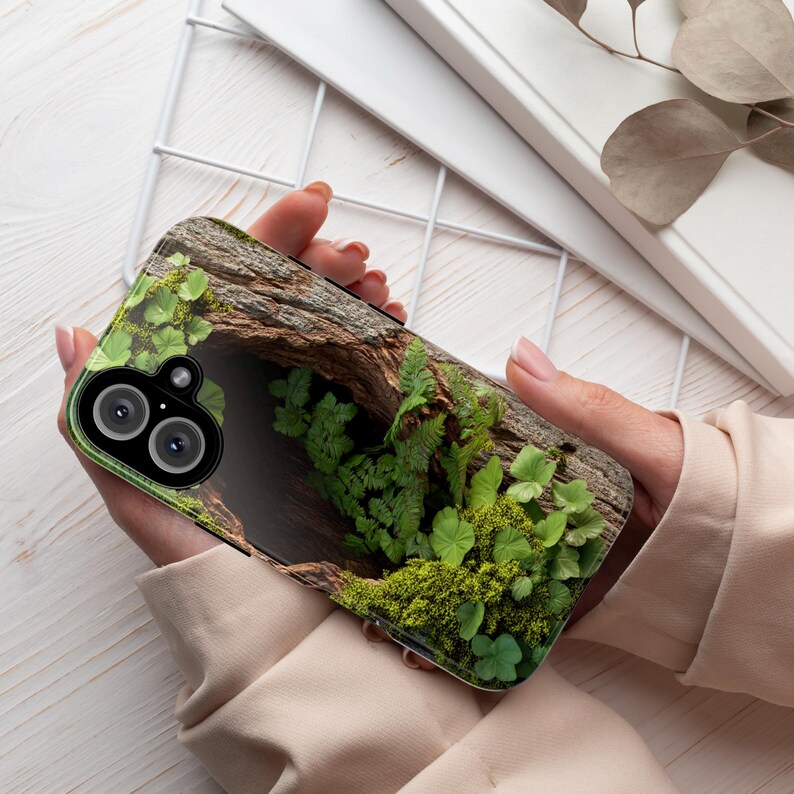 May include: A phone case with a green and brown nature scene. The case features a close-up of a tree trunk with moss and ferns growing around it.