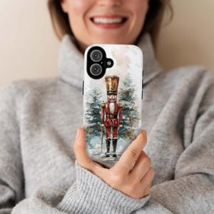 Puede incluir: Una funda de teléfono blanca con un diseño de acuarela de un cascanueces de pie en un bosque nevado. El cascanueces lleva un uniforme rojo y dorado y sostiene una espada.
