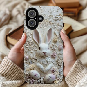 May include: White phone case featuring a 3D design of a white rabbit surrounded by flowers and leaves. The phone case has a black camera lens.