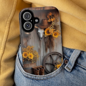 Puede incluir: Una funda transparente para teléfono con un diseño del oeste. La funda muestra un fondo de madera con una calavera de toro, sombrero de vaquero, girasoles y una rueda de carro. También se ven botas de vaquero marrones y un barril de madera.