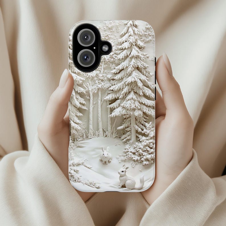 May include: A white phone case featuring a 3D design of a snowy woodland scene with two white rabbits. The case has a black camera lens cutout.