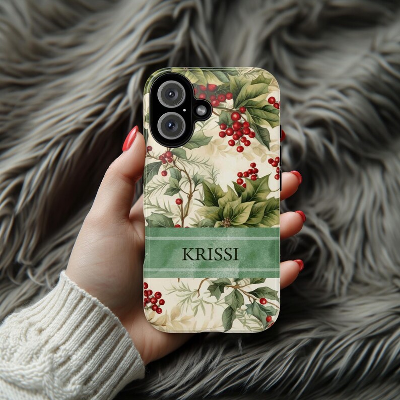 May include: A phone case featuring a botanical design with green leaves and red berries. The case has a cream-colored background and a green stripe with the name "KRISSI" in white. The case is held in a hand.