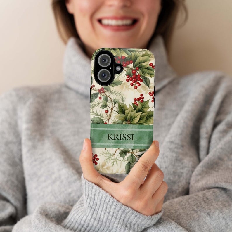May include: A phone case featuring a festive botanical design with holly leaves and red berries. A green banner at the bottom displays the name "KRISSI" in white letters. The case has a cream-colored background.
