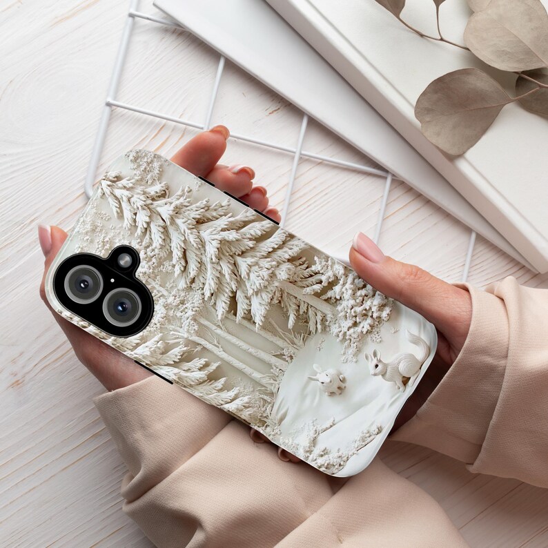May include: A white phone case featuring a winter forest scene. The case has a 3D-like design of snow-covered trees and two small white deer in the foreground.